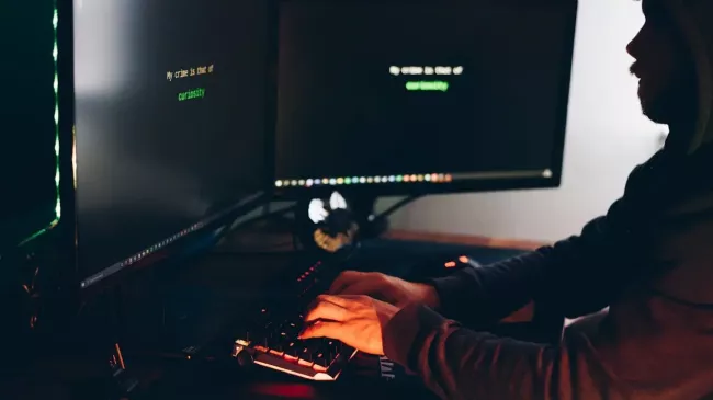 Hacker sedící v přítmí před dvěma monitory